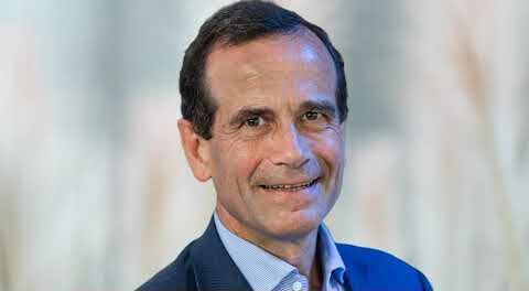 Portrait d’un homme souriant en costume bleu clair, devant un fond flou lumineux, représentant un cadre ou expert professionnel.