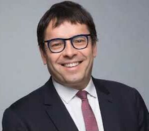 Portrait d’un homme souriant en costume foncé, cravate rouge et lunettes, illustrant un professionnel ou un cadre dirigeant
